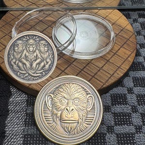 May include: Two bronze-colored coins with monkey designs. One coin features three monkeys, and the other has a single monkey face. Clear plastic coin holders are also visible, resting on a wooden surface.