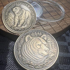 May include: Two bronze-colored coins with bear designs. One coin features a bear within a laurel wreath, the other depicts a bear's head with a forest backdrop. The coins are displayed near a clear plastic case.