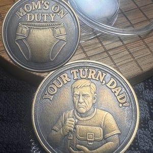May include: Two bronze-colored coins. One coin reads "MOM'S ON DUTY" with a diaper graphic. The other coin reads "YOUR TURN, DAD!" and features a man with a bag. The coins are on a wooden surface.