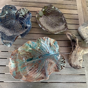 Puede incluir: Cuatro platos de cerámica con forma de hoja con un glaseado rústico. Los platos son en tonos de marrón, azul y verde. Un plato tiene una pequeña figura de rana en la parte superior.