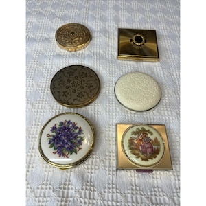 May include: A collection of six compact mirrors in various shapes and designs. Included are two round gold-tone metal compacts with floral engravings, one square gold-tone compact with a black center, one round white textured compact, one round compact with a purple floral needlepoint inlay, and one square gold-tone compact featuring a circular porcelain insert with a painted scene of two figures.