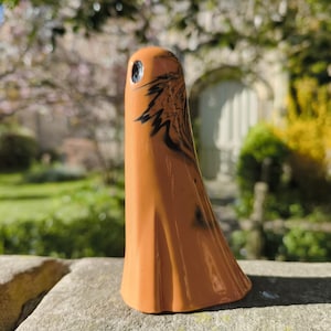 May include: An orange ceramic ghost ornament with a black design. The ghost has a small hole near the top. It stands on a stone surface, with a blurred outdoor scene in the background. The figurine is approximately 15 cm tall.