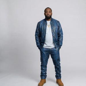 Peut inclure: Un ensemble veste bomber et pantalon bleu à motif peau de serpent. La veste a un col et des poignets côtelés. La tenue est associée à un t-shirt blanc et des bottes de travail beiges. Un collier chaîne en or est visible.