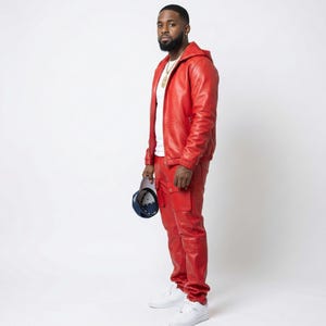 K&ouml;nnte beinhalten: Ein rotes Leder-Outfit, bestehend aus einer Jacke mit Kapuze und einer passenden Hose. Die Jacke hat einen Rei&szlig;verschluss und die Hose Cargotaschen. Die Person tr&auml;gt ein wei&szlig;es T-Shirt und wei&szlig;e Turnschuhe. Eine marineblaue und wei&szlig;e Baseballkappe wird in der Hand gehalten.