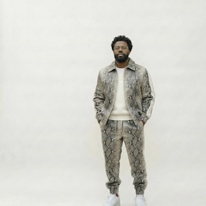 May include: A matching snakeskin print jacket and pants set. The jacket has a collared neckline and white stripe down the sleeves. The pants are joggers. The outfit is paired with white sneakers.