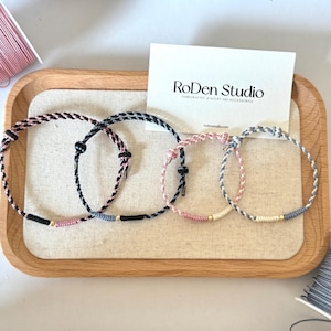 May include: Four braided cord bracelets in black, pink, and gray hues, each with a small gold bead accent. The bracelets are displayed on a wooden tray with a card that reads "RoDen Studio".