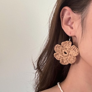 May include: A close-up of a woven, beige flower earring. The earring is a large, five-petal design with a circular center. The earring is suspended from a silver hook. The background is a blurred view of a person's hair and skin.