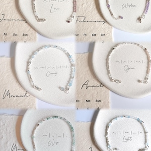 May include: Six beaded bracelets with silver clasps are displayed on white, heart-shaped dishes. Each bracelet features a different color of beads and a word in Morse code. The words are Love, Wisdom, Courage, Grace, Wise, and Light.