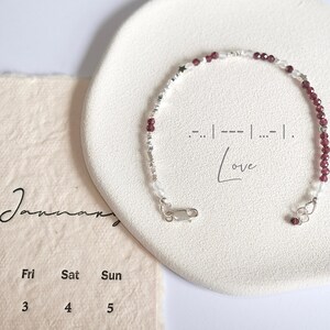 Könnte beinhalten: Ein silbernes Armband mit roten und weißen Perlen und einem Stern-Anhänger. Das Armband hat einen Verschluss und der Text '.-..|---|...|. Love' ist auf einer weißen Oberfläche hinter dem Armband geschrieben.