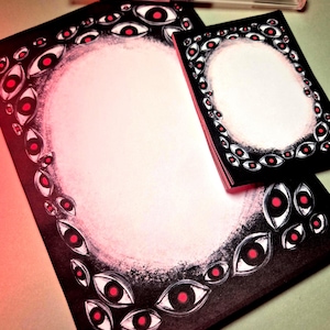 May include: A stationery set with a black and white eye design and red pupils. The set includes a notepad and notecards. A clear pen with red ink is placed on top of the stationery. The stationery is designed with a spooky theme.