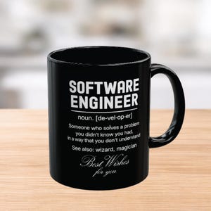Puede incluir: Taza de cerámica negra con texto blanco "SOFTWARE ENGINEER". La taza incluye una definición y las palabras "Best Wishes for you". La taza está sobre una superficie de madera.