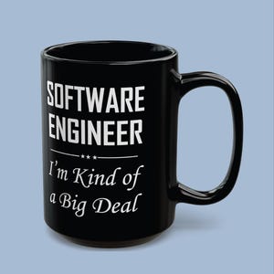 May include: A black ceramic mug with a large handle featuring white text that reads SOFTWARE ENGINEER followed by three small stars and the phrase I'm Kind of a Big Deal in a cursive font. The mug has a smooth, glossy finish and a cylindrical shape.