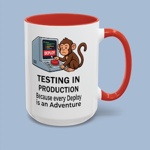 May include: A white ceramic mug with a red interior and handle. The front features a cartoon monkey pressing a red button on a computer console. Text reads: TESTING IN PRODUCTION Because every Deploy is an Adventure. The design is colorful and clear.
