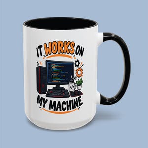Könnte beinhalten: Ein weißer Keramikbecher mit schwarzem Henkel und Rand, verziert mit einer Grafik eines Computer-Setups mit Monitor, Tastatur, Maus und einer kleinen Pflanze. Der Text "IT WORKS ON MY MACHINE" steht in schwarzen und orangen Buchstaben.