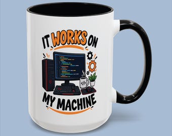 May include: A white ceramic mug with a black handle and rim, featuring a graphic of a computer setup with a monitor, keyboard, mouse, and a small plant. The text "IT WORKS ON MY MACHINE" is printed in black and orange lettering above and below.