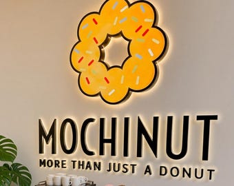 May include: A wall-mounted yellow donut sign with colorful sprinkles above black lettering reading MOCHINUT MORE THAN JUST A DONUT. Below, a silver espresso machine sits on a wooden table next to a three-tiered display stand holding twelve assorted donuts.