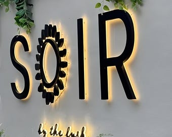 May include: A wall-mounted sign featuring the black letters S, O, I, R, with a black sun-shaped icon replacing the letter O. The sign is backlit with a warm yellow glow. Below, the text reads by the lush in a smaller, illuminated script font.