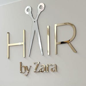 May include: A wall-mounted sign for a hair salon, featuring the word "HAIR" in gold-colored, three-dimensional letters, with a silver pair of scissors. The text "by Zara" is below the word "HAIR".