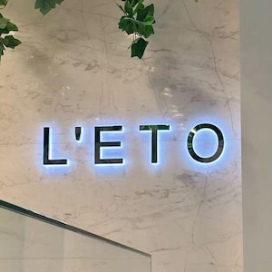 Puede incluir: Un letrero de pared con el texto iluminado L'ETO en letras oscuras y negritas sobre una superficie de m&aacute;rmol blanco con vetas grises. Follaje artificial de hiedra verde cuelga del techo sobre el letrero, y una vitrina de cristal se ve debajo.