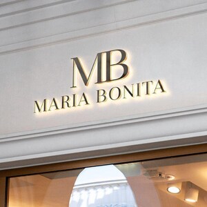 Puede incluir: Un letrero de pared tridimensional que presenta las letras MB en una fuente serif grande sobre el texto MARIA BONITA en una fuente serif m&aacute;s peque&ntilde;a. Las letras oscuras est&aacute;n montadas en una pared clara con un efecto de luz de fondo.