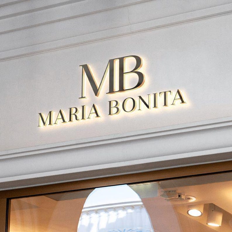 Puede incluir: Un letrero de pared tridimensional que presenta las letras MB en una fuente serif grande sobre el texto MARIA BONITA en una fuente serif m&aacute;s peque&ntilde;a. Las letras oscuras est&aacute;n montadas en una pared clara con un efecto de luz de fondo.