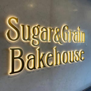 Puede incluir: Un letrero de pared con el texto dorado Sugar & Grain Bakehouse en una fuente con serifa. Las letras individuales est&aacute;n retroiluminadas con un brillo blanco c&aacute;lido, creando un efecto de halo sobre la pared gris claro de aspecto marmolado.