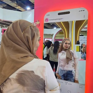 Op de afbeelding: Een grote, rechthoekige spiegel ontworpen als een smartphone-interface met een social media-profiel. Het frame bevat een digitale klok op 12:00 uur, de profielnaam koreashopabcegypt en interactieve iconen voor likes en reacties.