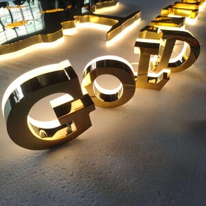 Puede incluir: Letras de metal de color dorado en tres dimensiones que forman la palabra GOLD, con una superficie pulida y reflectante e iluminaci&oacute;n LED trasera de color blanco c&aacute;lido que proyecta un suave resplandor sobre la superficie blanca.