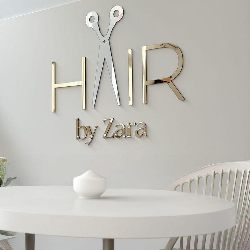 May include: A wall-mounted sign for a hair salon, featuring the word "HAIR" in gold-colored, three-dimensional letters, with a silver pair of scissors. The text "by Zara" is below the word "HAIR".