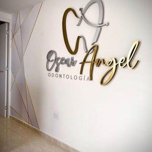 May include: A wall-mounted sign for "Oscar Angel Odontolog&iacute;a" in a dental office. The sign features a stylized tooth and the name in gold and silver lettering. The wall is white with geometric gold accents.