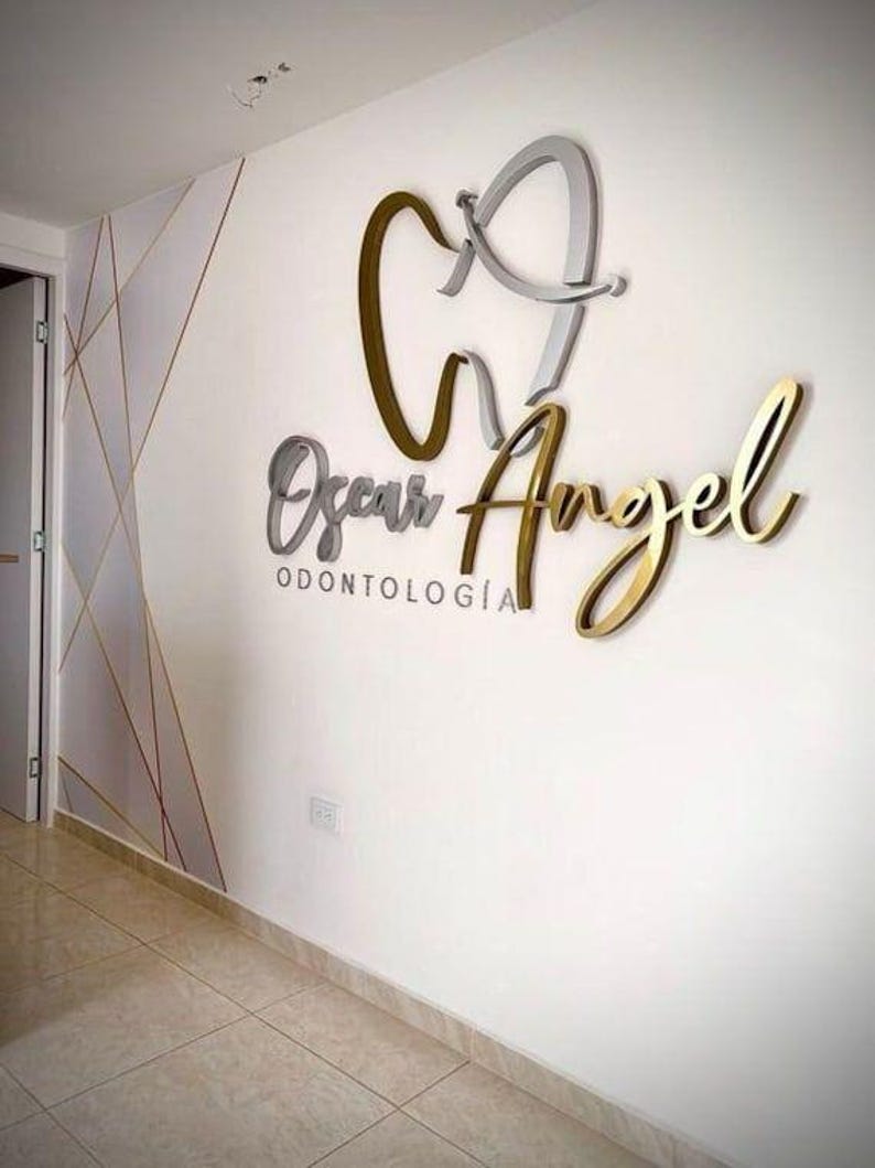 May include: A wall-mounted sign for "Oscar Angel Odontolog&iacute;a" in a dental office. The sign features a stylized tooth and the name in gold and silver lettering. The wall is white with geometric gold accents.
