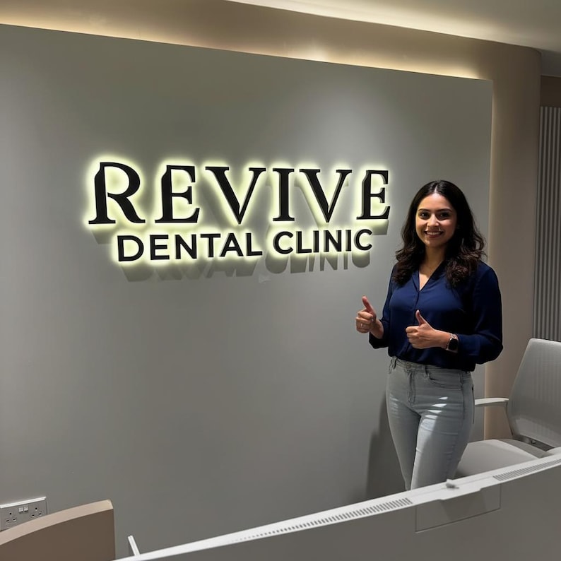 Puede incluir: Un letrero de pared con letras retroiluminadas que dice REVIVE DENTAL CLINIC en una fuente serif oscura. El letrero est&aacute; montado sobre una pared lisa de color gris claro, creando un suave resplandor detr&aacute;s de las letras. El dise&ntilde;o es limpio.
