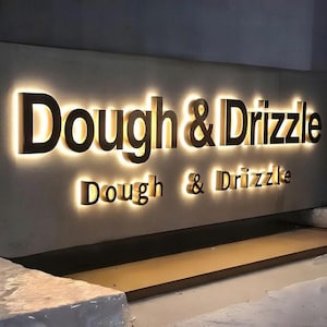 Peut inclure: Enseigne lumineuse pour "Dough & Drizzle". Le signe présente des lettres de couleur bronze avec une lueur dorée et chaleureuse. Le texte est affiché en grand au-dessus d'une version plus petite, probablement pour une boulangerie ou un restaurant.