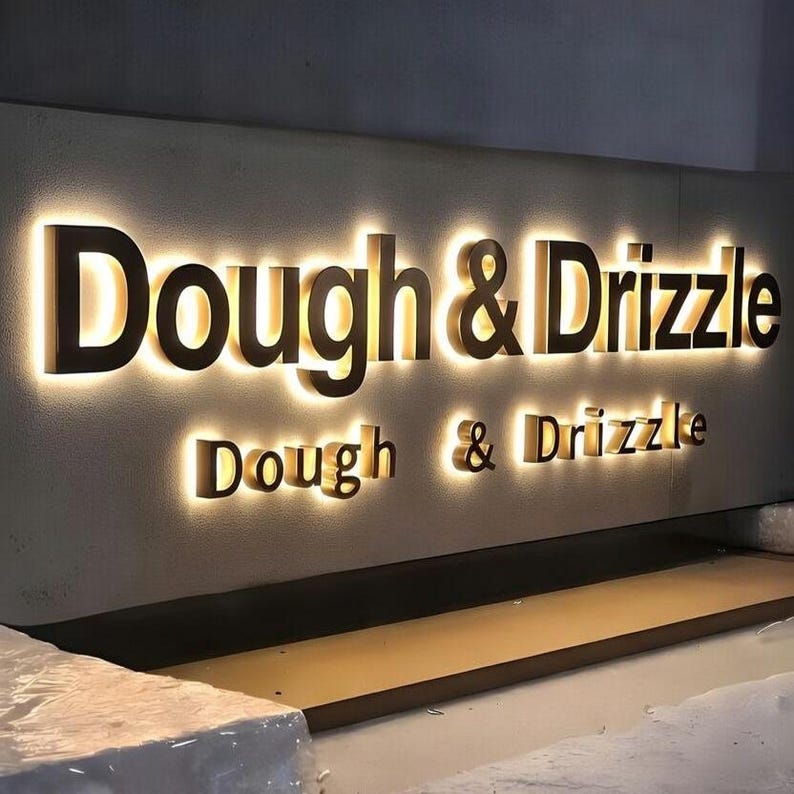 Puede incluir: Un letrero de pared con el texto Dough & Drizzle en letras grandes, oscuras y tridimensionales. Las letras est&aacute;n retroiluminadas con una luz c&aacute;lida y brillante que crea un efecto de halo sobre la superficie texturizada y clara de fondo.