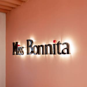 Puede incluir: Un letrero de pared con letras negras tridimensionales que forman el texto "Miss Bonnita" sobre una pared de color melocot&oacute;n. Las letras est&aacute;n retroiluminadas, proyectando un brillo c&aacute;lido sobre la superficie, con un peque&ntilde;o acento circular rojo sobre la i.