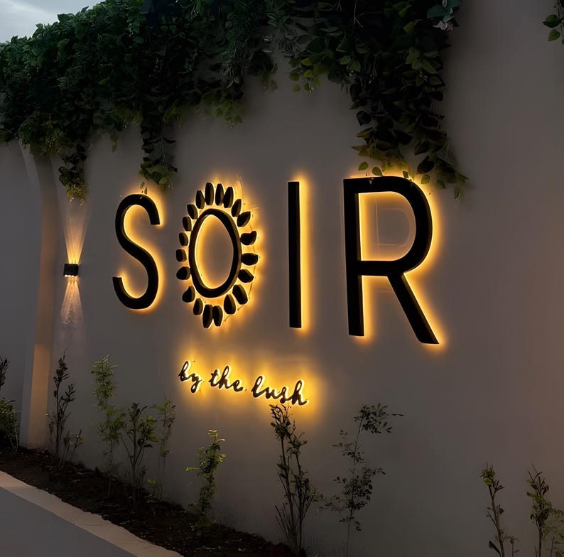 Puede incluir: Un letrero de pared con las letras negras S, O, I y R, con un icono en forma de sol que reemplaza la letra O. El letrero est&aacute; retroiluminado con un brillo c&aacute;lido, y el texto "by the lush" est&aacute; escrito debajo en una fuente cursiva negra.