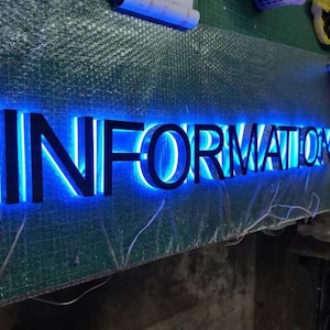 Puede incluir: Un letrero personalizado con la palabra INFORMATION en letras may&uacute;sculas negras, negritas y tridimensionales. El letrero est&aacute; iluminado desde atr&aacute;s con un brillo LED azul brillante que delinea cada car&aacute;cter, creando un efecto de halo.