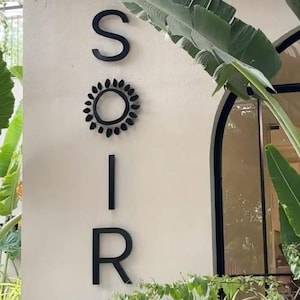 Puede incluir: Un letrero de pared vertical con las letras negras S, I y R, con un icono de sol circular negro que reemplaza la letra O. La palabra SOIR se muestra en una fuente sans-serif limpia sobre una pared de color claro, rodeada de follaje verde.