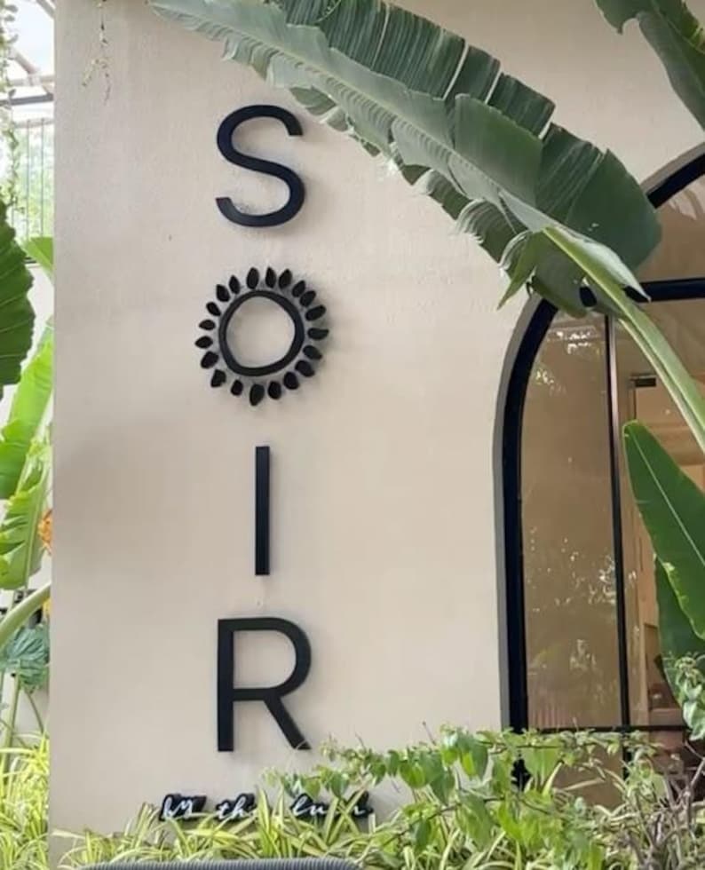 Puede incluir: Un letrero de pared vertical con las letras negras S, I y R, con un icono de sol circular negro que reemplaza la letra O. La palabra SOIR se muestra en una fuente sans-serif limpia sobre una pared de color claro, rodeada de follaje verde.