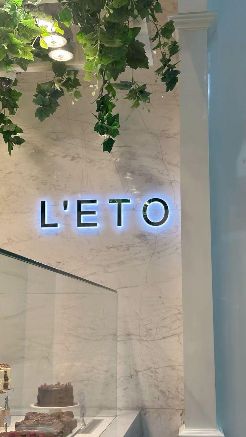 Puede incluir: Un letrero de pared con el texto iluminado L'ETO en letras oscuras y negritas sobre una superficie de m&aacute;rmol blanco con vetas grises. Follaje artificial de hiedra verde cuelga del techo sobre el letrero, y una vitrina de cristal se ve debajo.