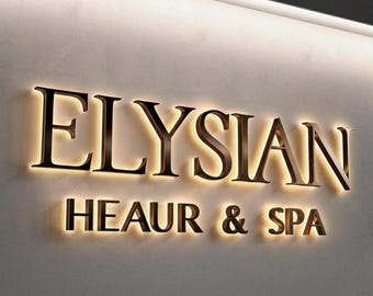 Op de afbeelding: Een aan de muur bevestigd bord met het woord "ELYSIAN" in grote, bronskleurige letters boven de woorden "HEAUR & SPA". De letters worden van achteren verlicht, waardoor een warme, uitnodigende gloed ontstaat. Het bord is op een lichtgekleurde muur gemonteerd.