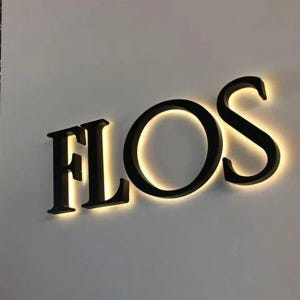 Puede incluir: Un letrero de pared tridimensional con las letras may&uacute;sculas negras F, L, O y S. Cada letra est&aacute; montada ligeramente separada de la pared, creando un efecto de halo brillante con luz blanca c&aacute;lida que emana detr&aacute;s de los bordes de los caracteres.