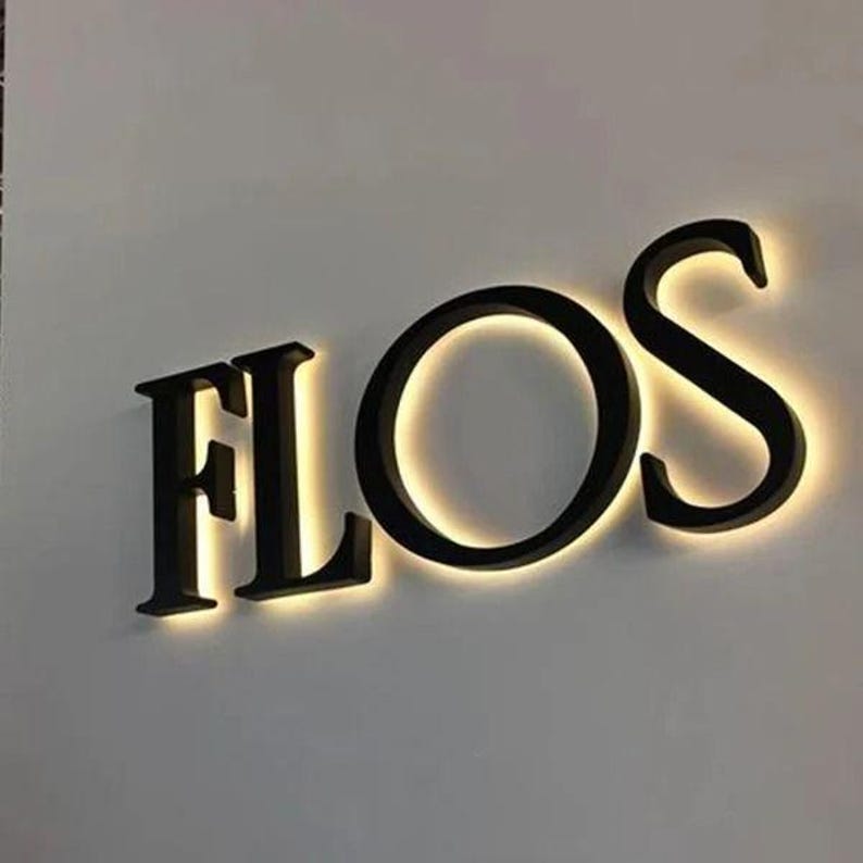 Puede incluir: Un letrero de pared tridimensional con las letras may&uacute;sculas negras F, L, O y S. Cada letra est&aacute; montada ligeramente separada de la pared, creando un efecto de halo brillante con luz blanca c&aacute;lida que emana detr&aacute;s de los bordes de los caracteres.