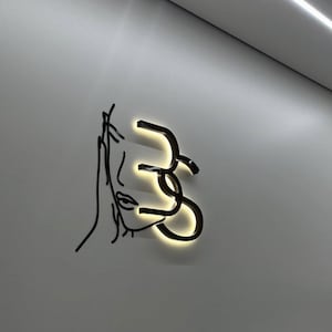 Puede incluir: Una pieza de arte mural de metal negro que representa el rostro de una mujer y formas abstractas. El arte está iluminado con una luz cálida, creando un contraste llamativo con la pared gris. El diseño es minimalista y moderno.