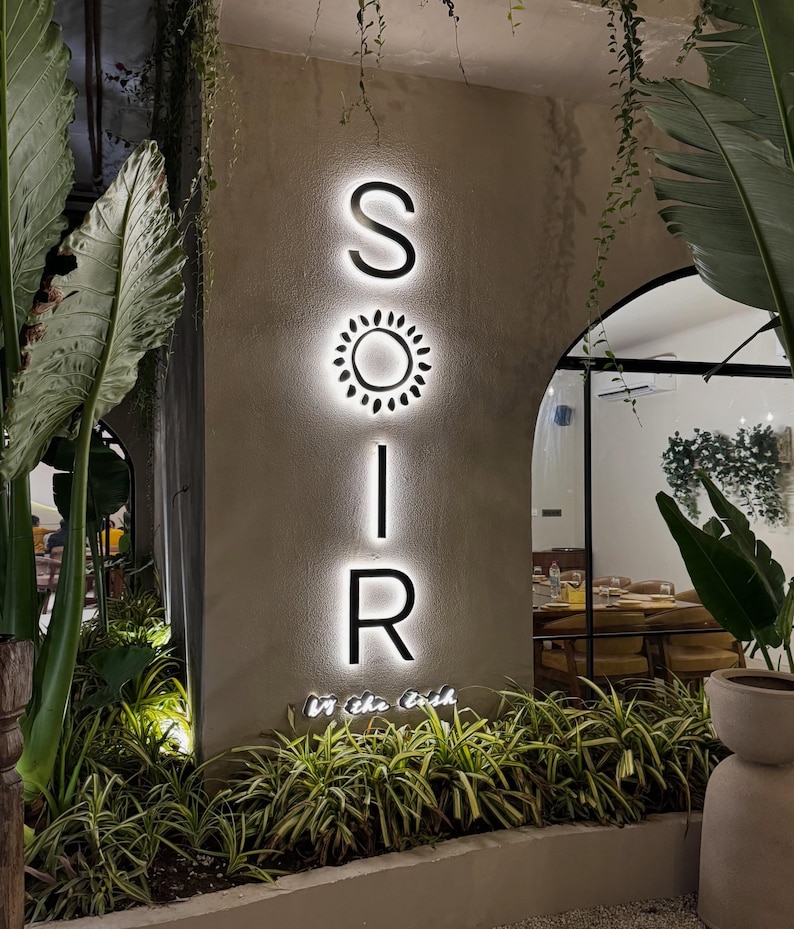 Puede incluir: Un letrero de pared vertical con las letras retroiluminadas S, O, I, R, donde un icono de sol reemplaza la letra O. El letrero est&aacute; montado en una pared beige texturizada, iluminado desde atr&aacute;s y rodeado de exuberantes plantas tropicales.