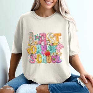 May include: Cream-colored t-shirt with a colorful graphic that reads "LAST DAY OF SCHOOL." The design includes an apple, glasses, a star, and a pencil. The shirt is worn by a person sitting in a white chair.