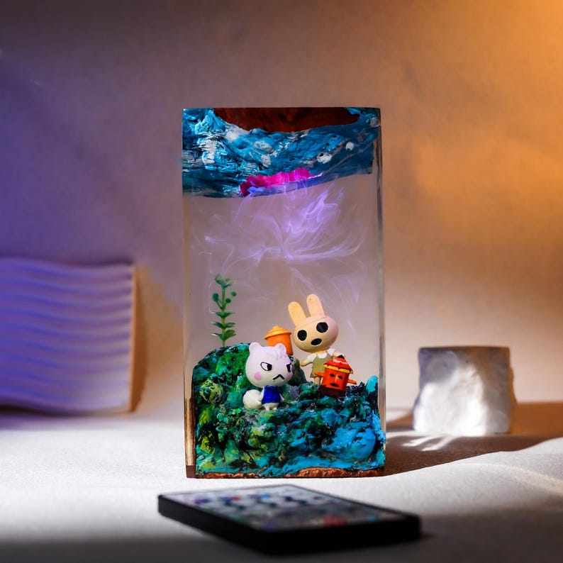 Może przedstawiać: Prostokątny element artystyczny z żywicy z miniaturową dioramą. Diorama zawiera niebiesko-zielony krajobraz z dwiema postaciami z kresk&oacute;wek, białym kotem i brązowym kr&oacute;likiem. G&oacute;rna część elementu ma niebieskie i białe wiry, przypominające wodę.