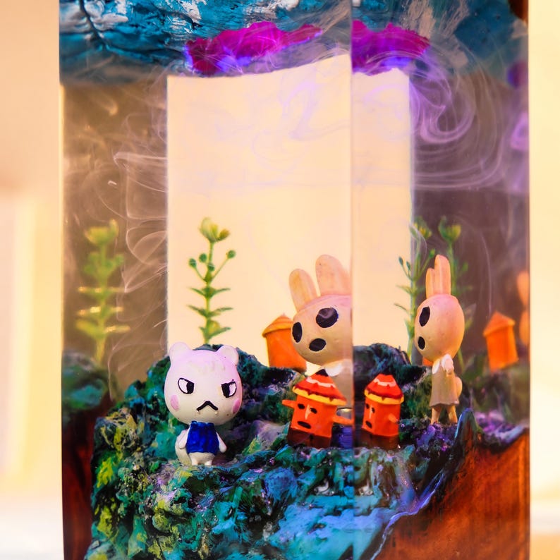 Może przedstawiać: Dekoracyjne dzieło sztuki z żywicy z miniaturowymi figurkami postaci z kresk&oacute;wek w scenerii krajobrazu. Scena obejmuje białego kota, dwa kr&oacute;liki, małe pomarańczowe konstrukcje i zielone liście, wszystko na tle niebieskiej i białej żywicy.