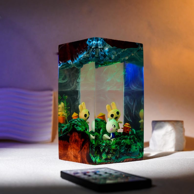 Może przedstawiać: Prostokątny element artystyczny z żywicy przedstawiający miniaturową dioramę z postaciami zwierząt z kresk&oacute;wek. Blok żywicy ma niebiesko-brązową kolorystykę, z żywą zieloną podstawą i ciemną g&oacute;rą. Smartfon znajduje się na pierwszym planie.