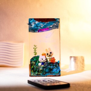 Może przedstawiać: Prostokątny element artystyczny z żywicy z miniaturową dioramą. Przedstawia postacie z kresk&oacute;wek, w tym białego kota i kr&oacute;lika, na niebiesko-zielonej podstawie. G&oacute;rna część ma abstrakcyjną niebieską i brązową sztukę.